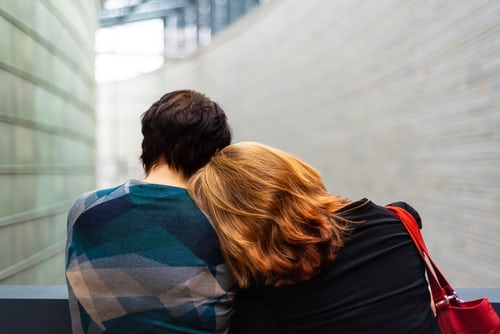 A couple facing away from you, the lady rests her head on her partners shoulder for comfort.t