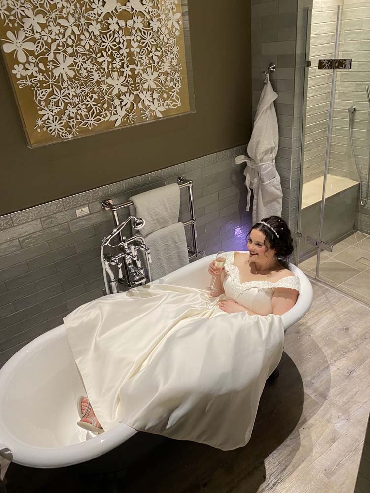 A bride in full wedding attire sitting in the bath of her bridal suite.