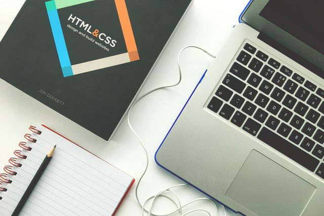 A web designer's desk with a laptop, notebook, and book about HTML