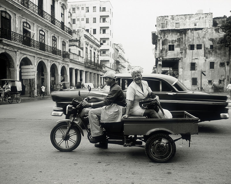 zwartwitfoto van een brommer met bak eraan vast op straat in Cuba met een toerist die vervoerd wordt