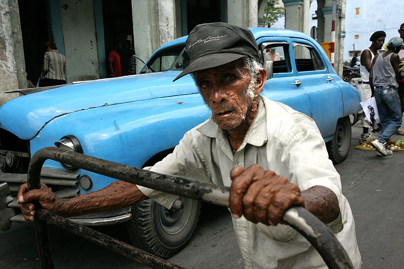 foto van man die kar vasthoudt in een drukke straat met blauwe auto op achtergrond n Cuba