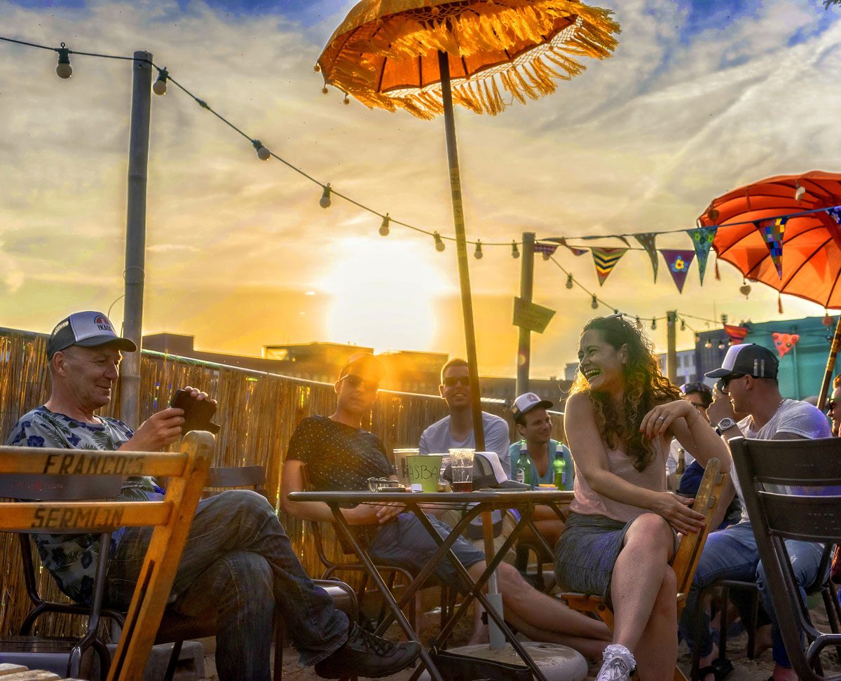 vrolijke foto van mensen op het terras van een strandtent met zon erachter die bijna ondergaat