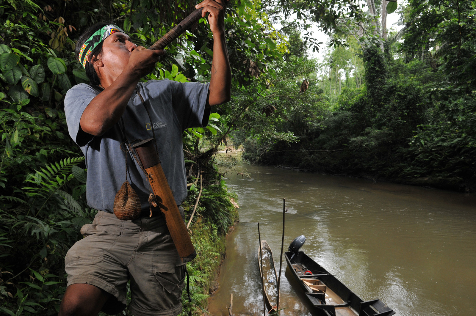 foto van een indiaan die bij een rivier met een boot in een bos blaast op een pijp die hij naar boven richt