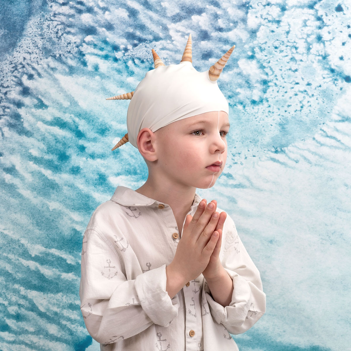foto van jongetje metr badmuts op met hanenkammen van schelpen zijn handen voor zijn borst gevouwen als namaste met achtergrond van een witte schuimende zee