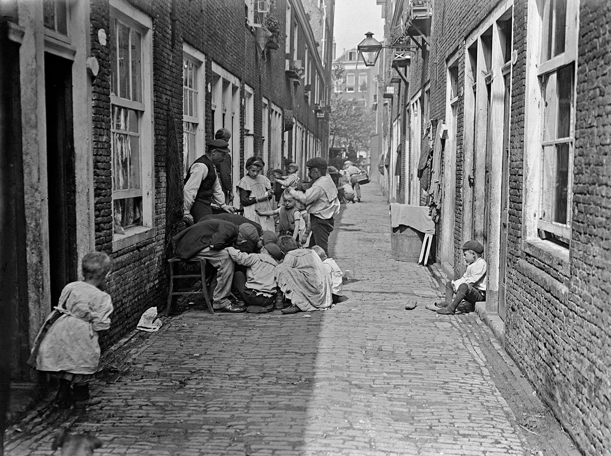 foto uit 1910 in zwartwit van een straatje in Rotterdam met mensen voor hun huis op stoel en staand en op grond