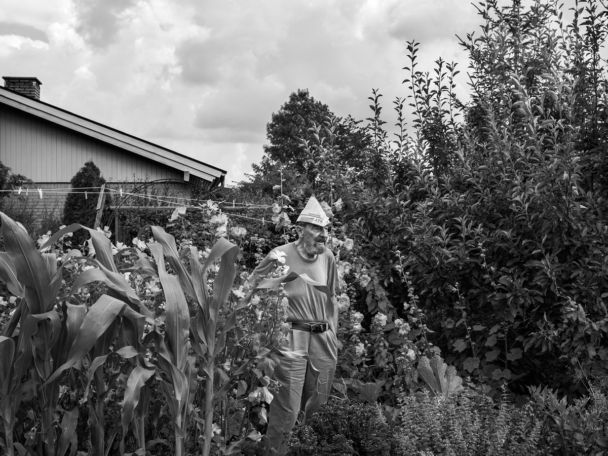 foto van een man met een papieren hoedje in tuin tussen hoge bloemen en struiken