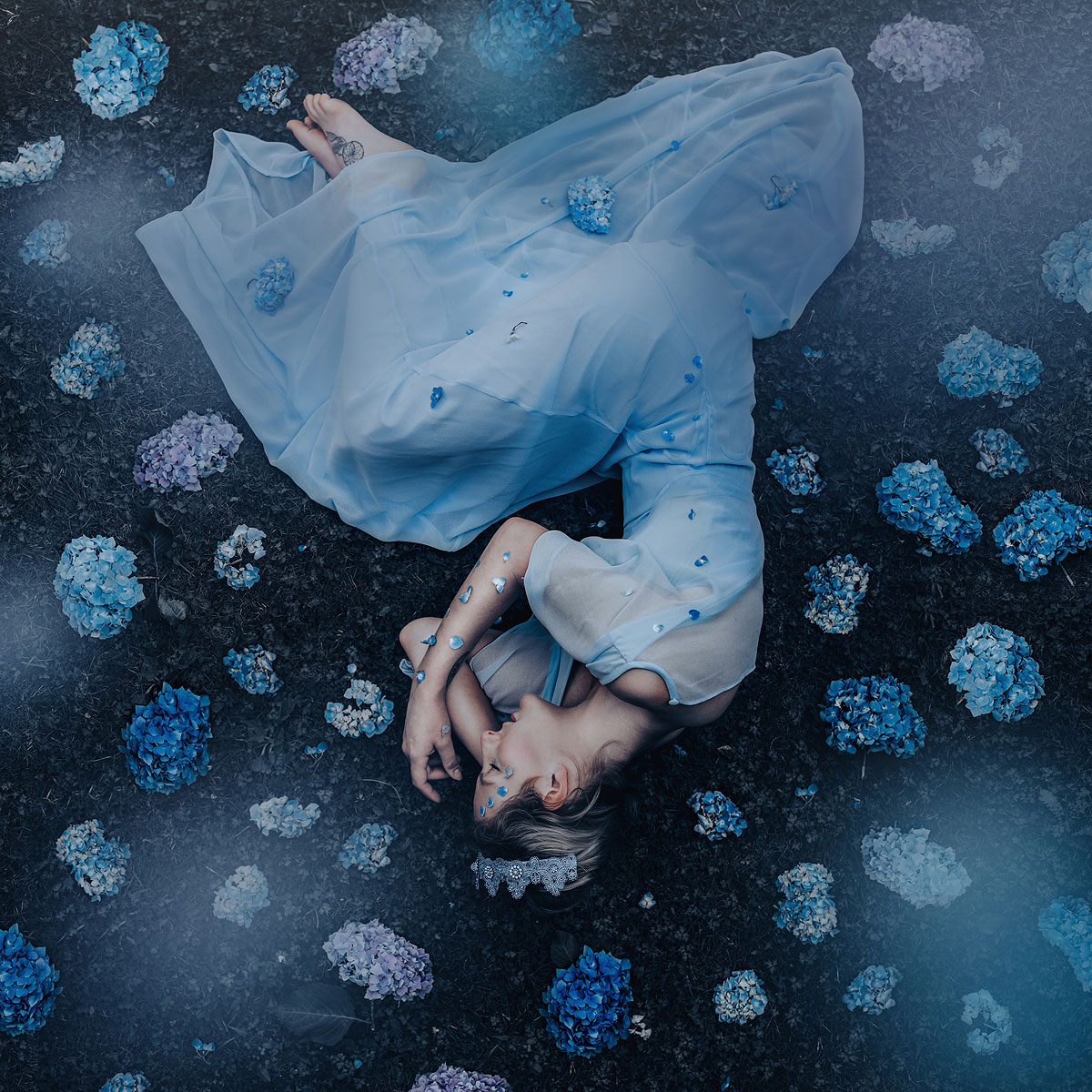 foto van een vrouw die op haar zij ligt, het hoofd onderaan, in een blauwe jurk met blauwe hortensia bloemen eromheen