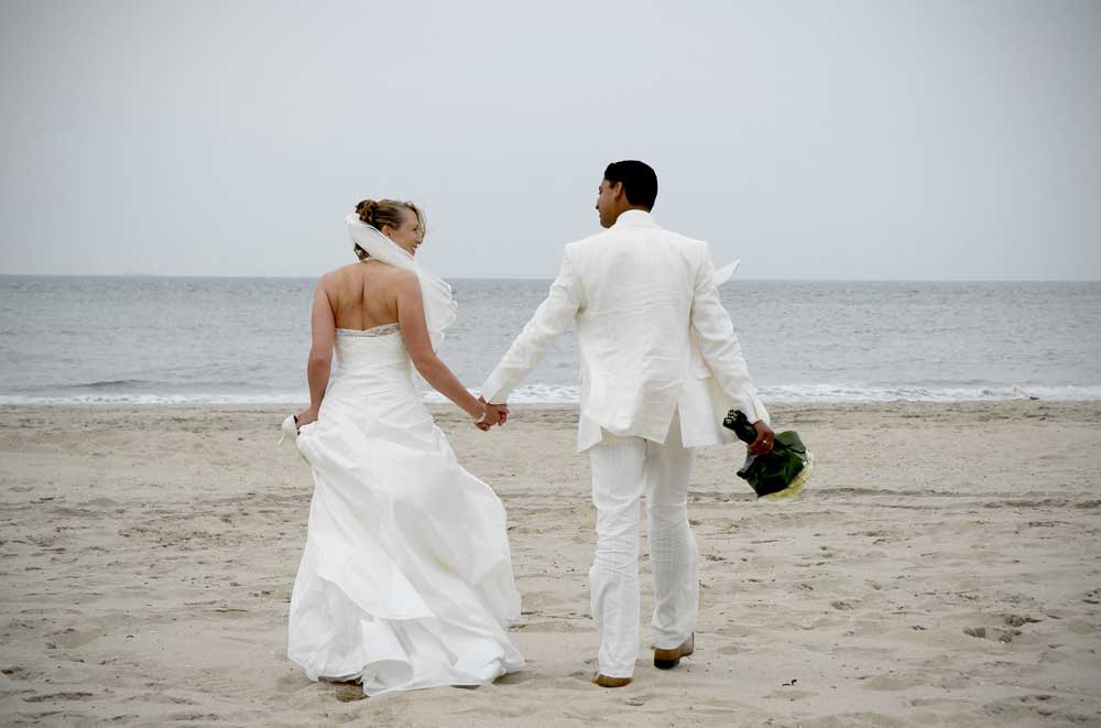 trouwfoto van echtpaar in wit op het strand hand in hand lopend naar de zee