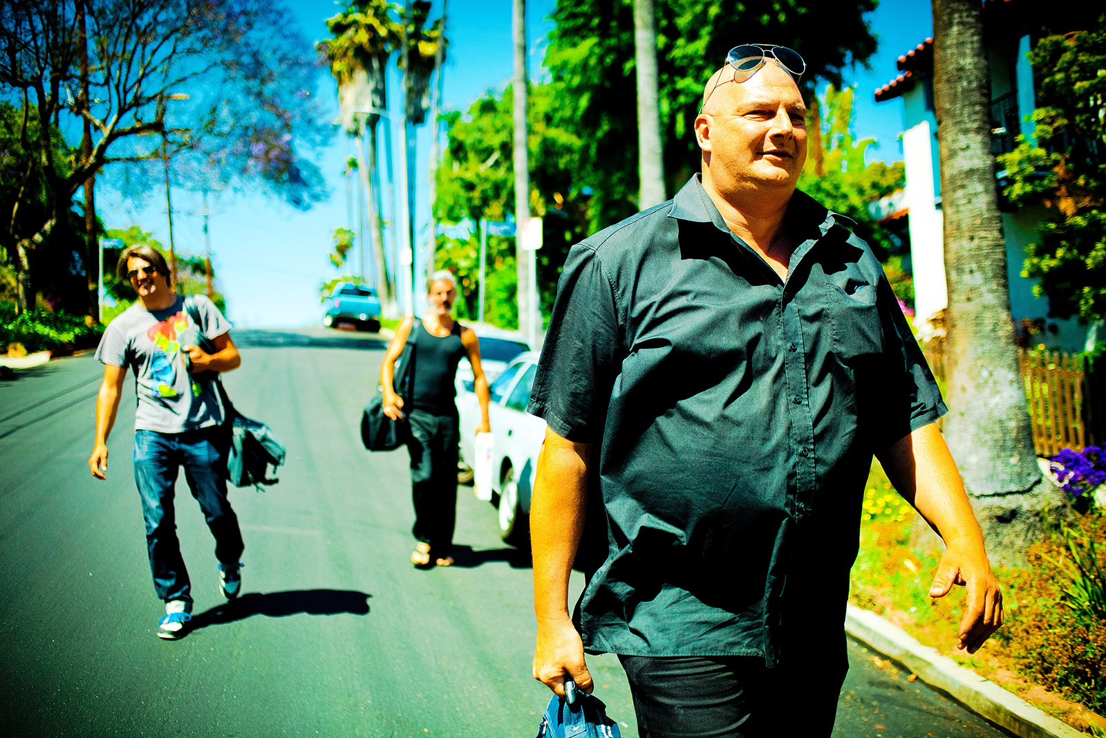 foto van drie mensen die op straat lopen in een zonnig land, een dikke man voorop en jongen links erachter en een andere man iets verder naar achter