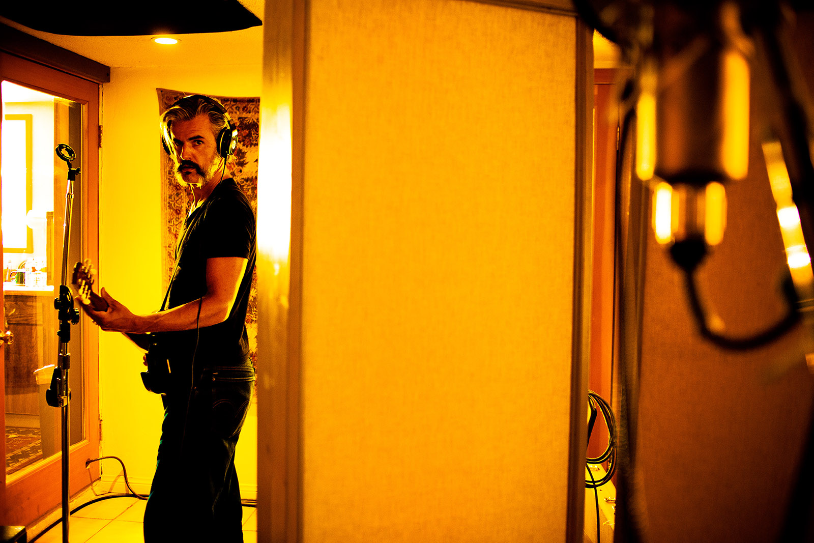 foto van een man met elektrisch gitaar en koptelefoon op in een kamer die gelig verlicht is