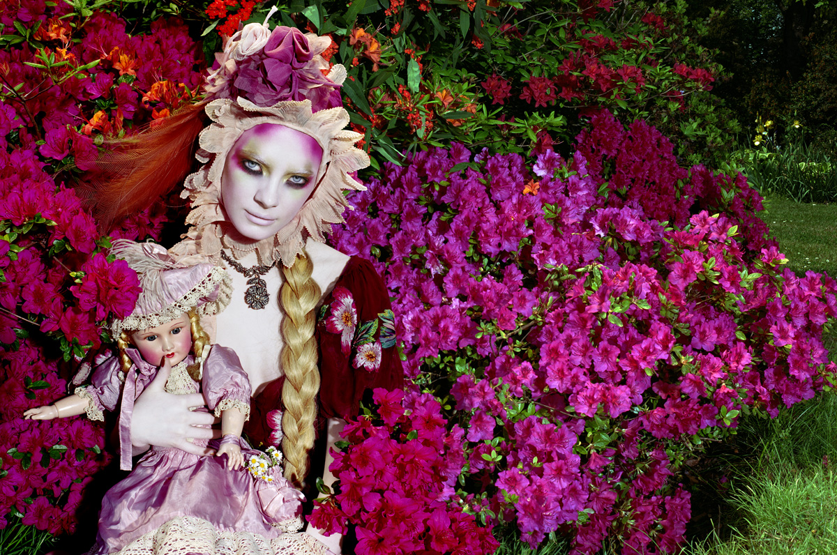 foto van Miles Aldridge van vrouw tussen bloemenstruiken met blonde vlecht en pop in arm en hoed van bloemen