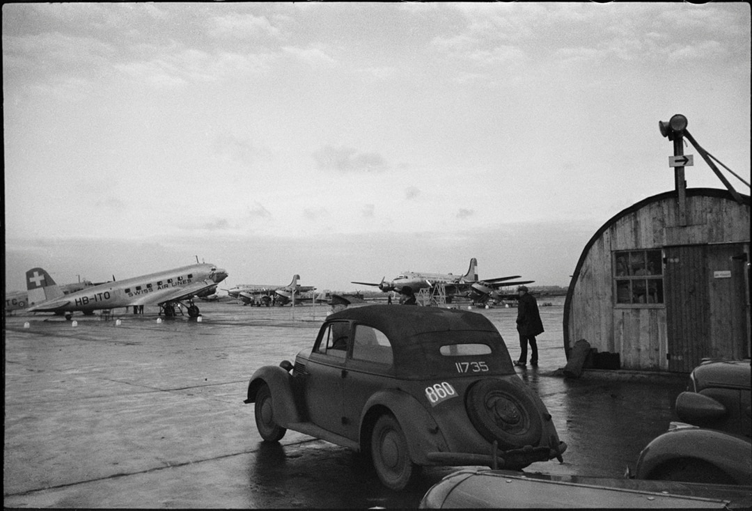 foto van werner bischof van een vliegtuigbasis in zwartwit met een deux cheveaux op voorgrond