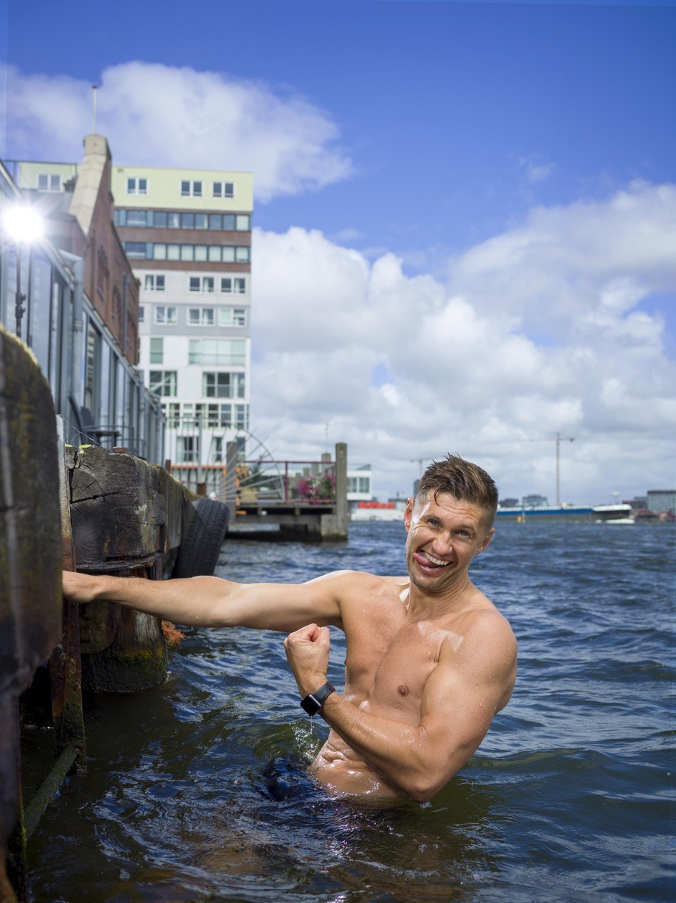 foto van man met ontbloot bovenlijf die zijn tong uitsteekt en een vuist maakt in water bij kade van rivier
