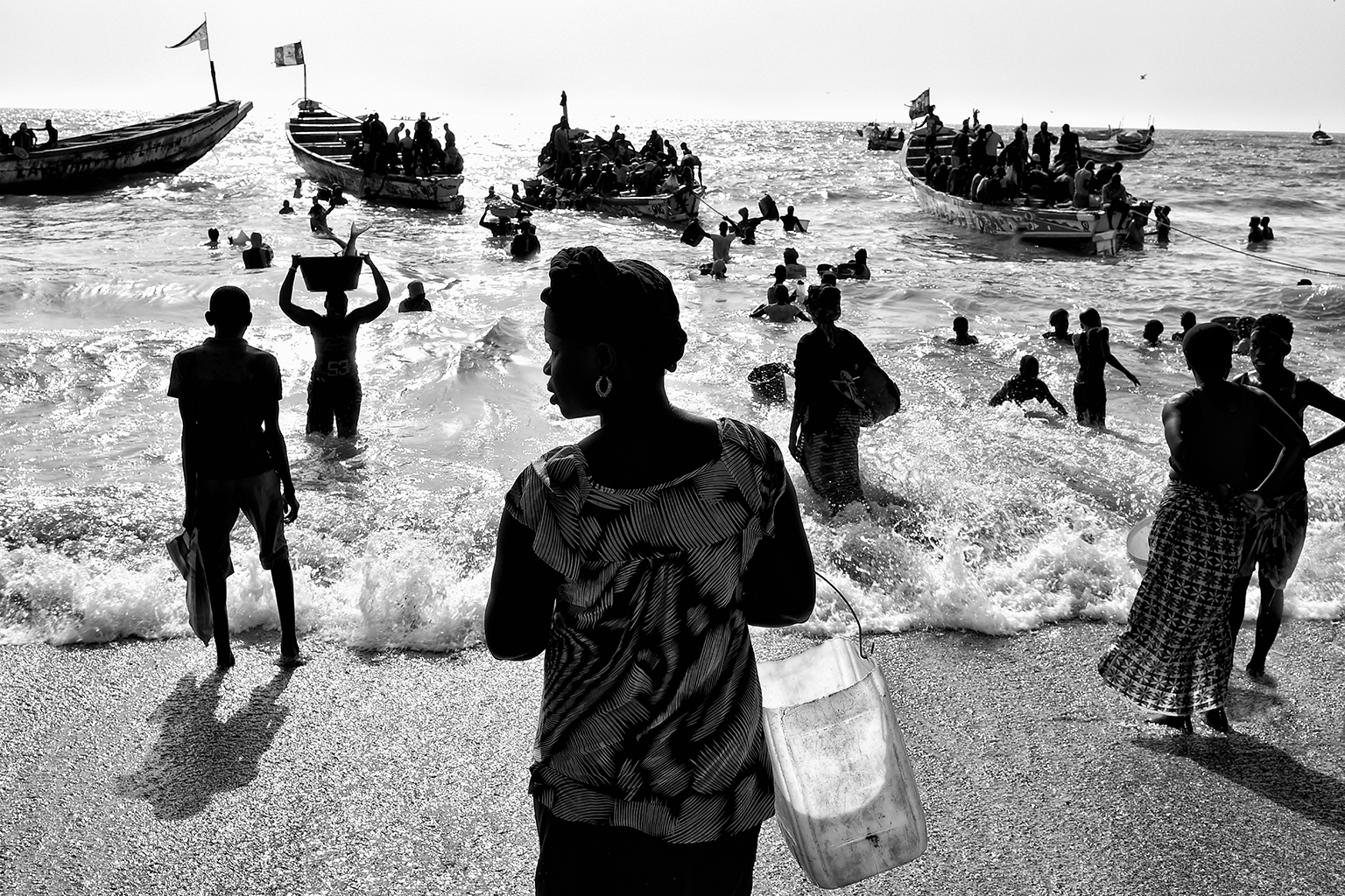 foto van strand waar de zee begint in Gambia in zwartwit waar allerlei mensen staan met manden en vissersboten