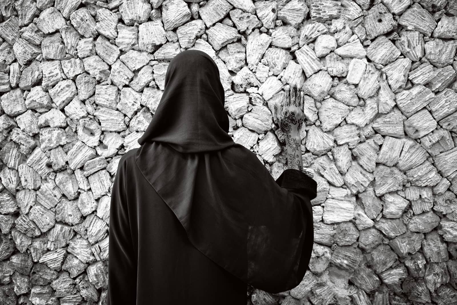 © Annelies Damen - Fikra, vrouw in zwart gekleed met zwart hoofddoek staat met rug naar camera tegen een muur met hand op muur