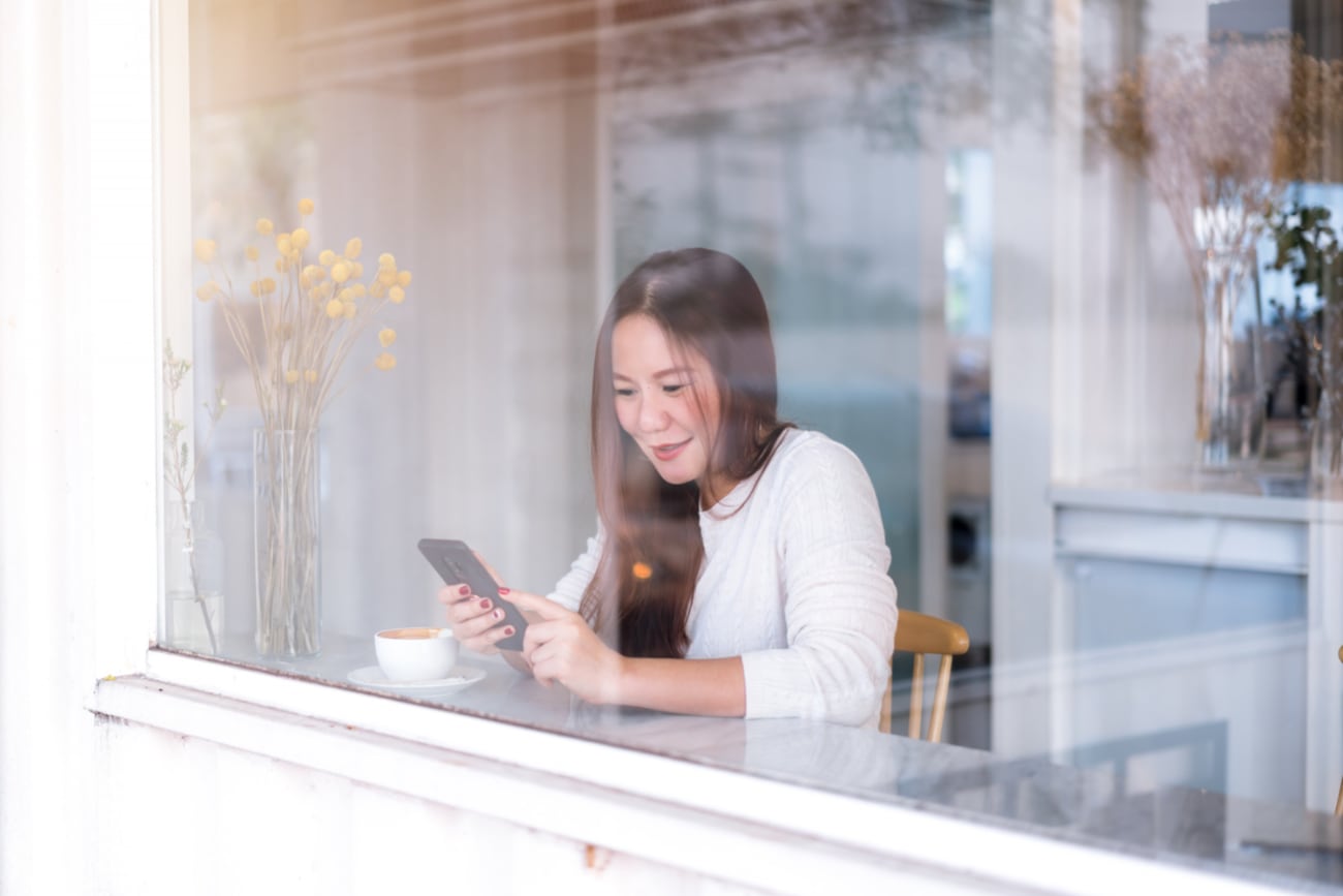 young-asian-woman-relax-using-phone-shot-through-glass-window-coffee-shop