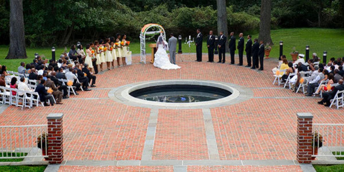 a bride and groom during their wedding ceremony. their friends are arranged in a circle around a fountain behind the happy couple on a red brick patio