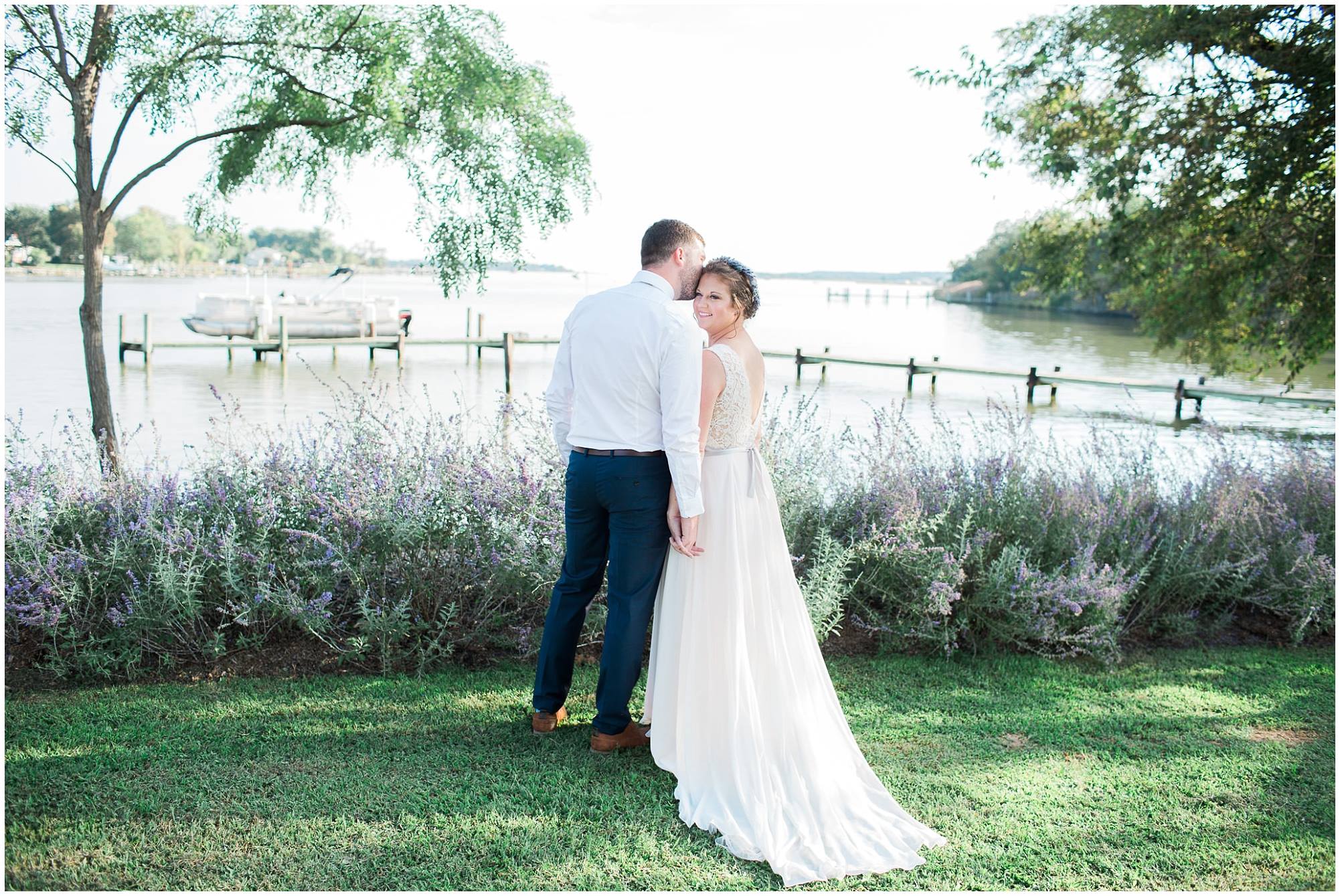 a bride and groom pose by a bay, the groom leaning over to kiss the bride and the bride looking back at the camera
