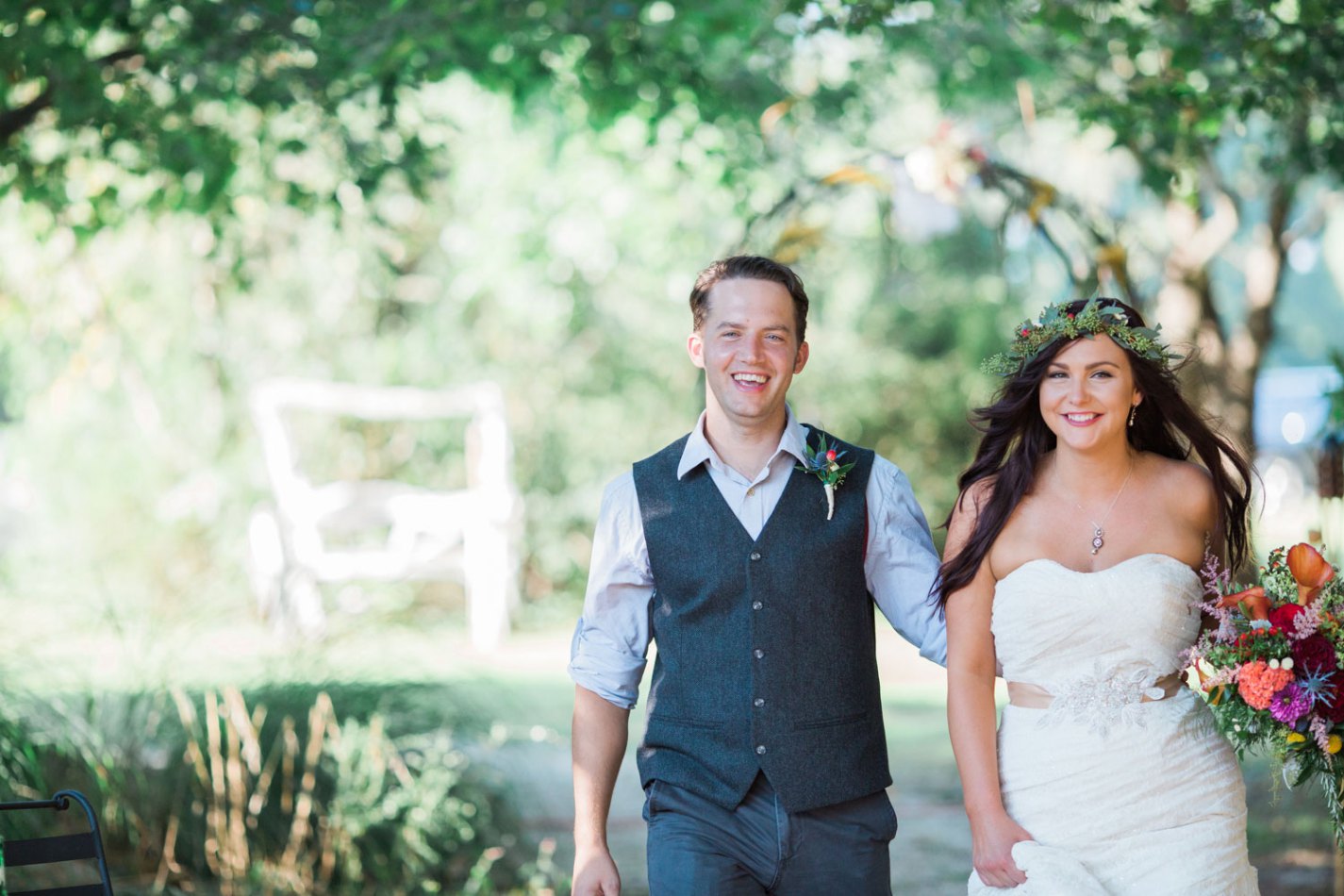a bride wearing a wreath of flowers on her head and carrying her bouquet walks with her new husband, who has taken his jacket off and rolled up his sleeves