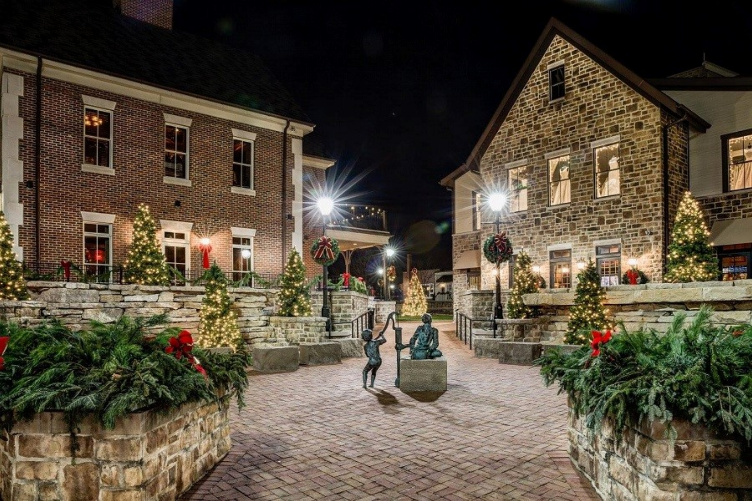 A cobblestone street with two historic brick buildings aglow with decorated christmas trees and planters full of red poinsettia flowers