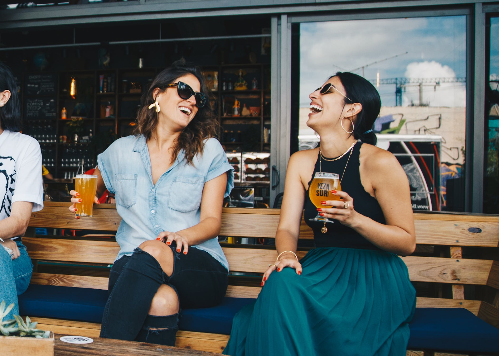 two female friends sit on a bench drinking and laughing at happy hour