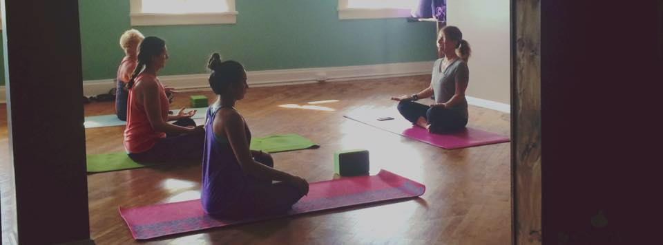 Photo of Yoga Class. Instructor is sitting the front in an upright position.