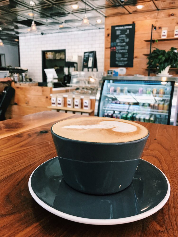latte filled to the rim of a coffee cup. background includes the coffee shop and case full of other drink items