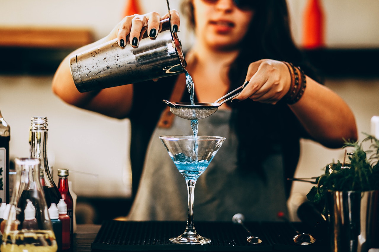 A bartender stands making a blue cocktail. In her hands are a spoon and a drink shaker.