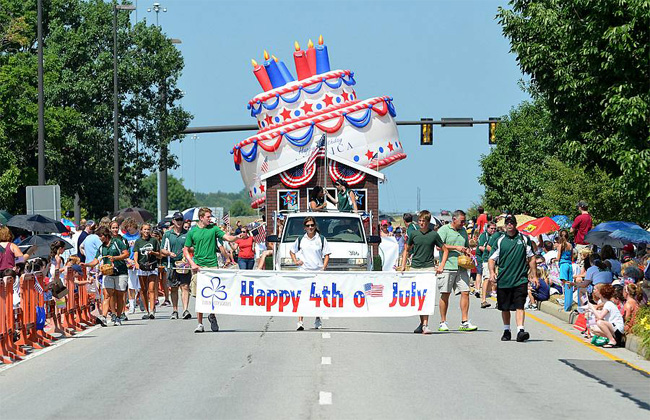 young men holding "Happy 4th of July" sign in front of large American flag-colored cake during a parade