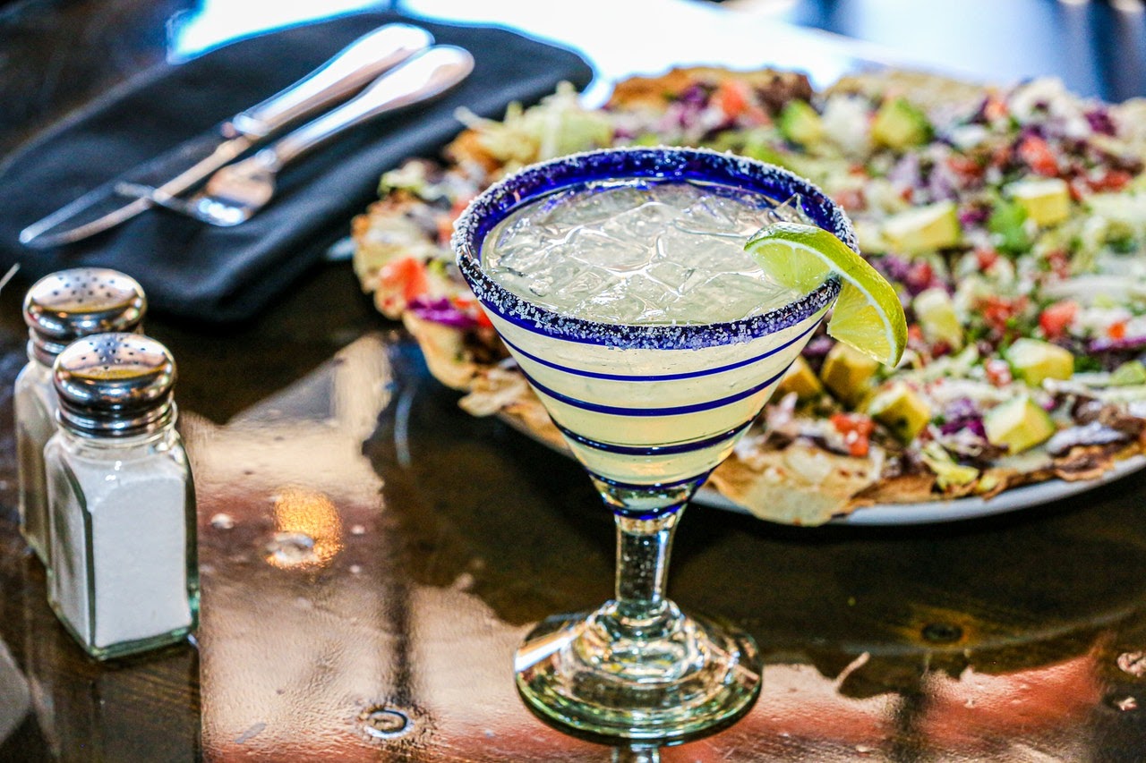 A closeup of a green margarita with salt and a lime on the rim. Behind it is a plate of loaded nachos..