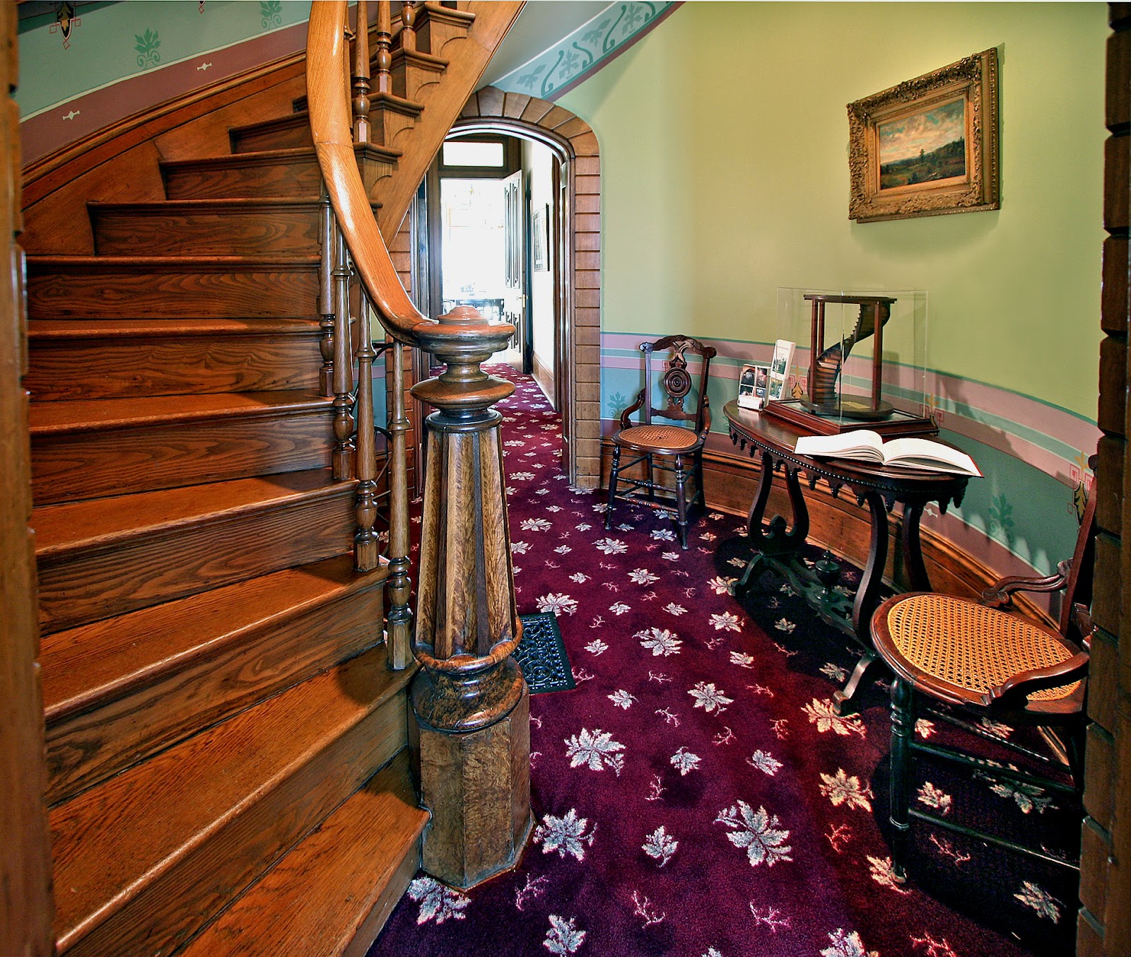 A wooden, curved stairway amid a patterned maroon carpet, green walls, and other beautiful old wooden pieces