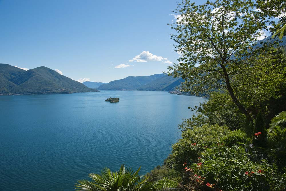 Krásně modré jezero Maggiore obklopené zelenými horami, rostlinami a stromy.