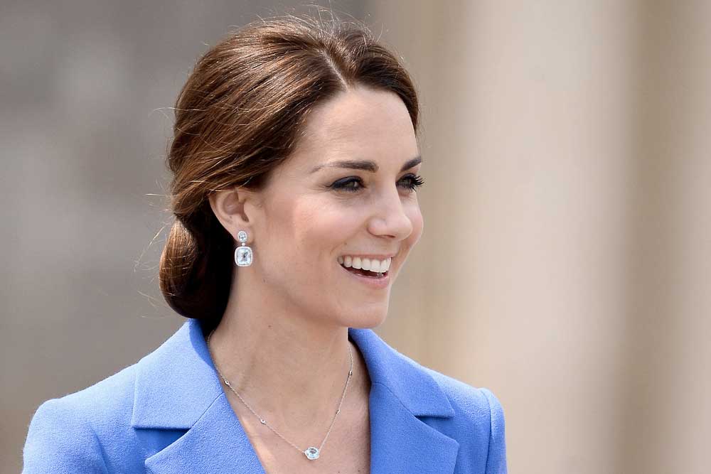 Okouzlující Kate Middleton v modrém kostýmku se s okouzlujícím úsměvem dívá do davu.