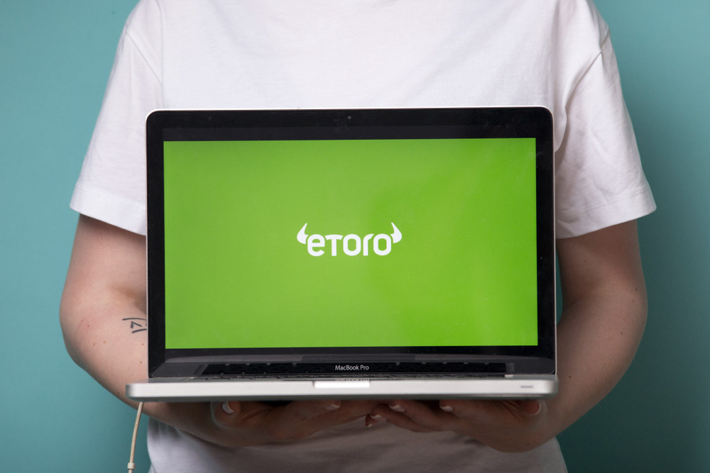 Mladík v bílém tričku, který drží notebook, na kterém se ukazuje logo firmy eToro. Ta patří mezi největší brokery na světě.