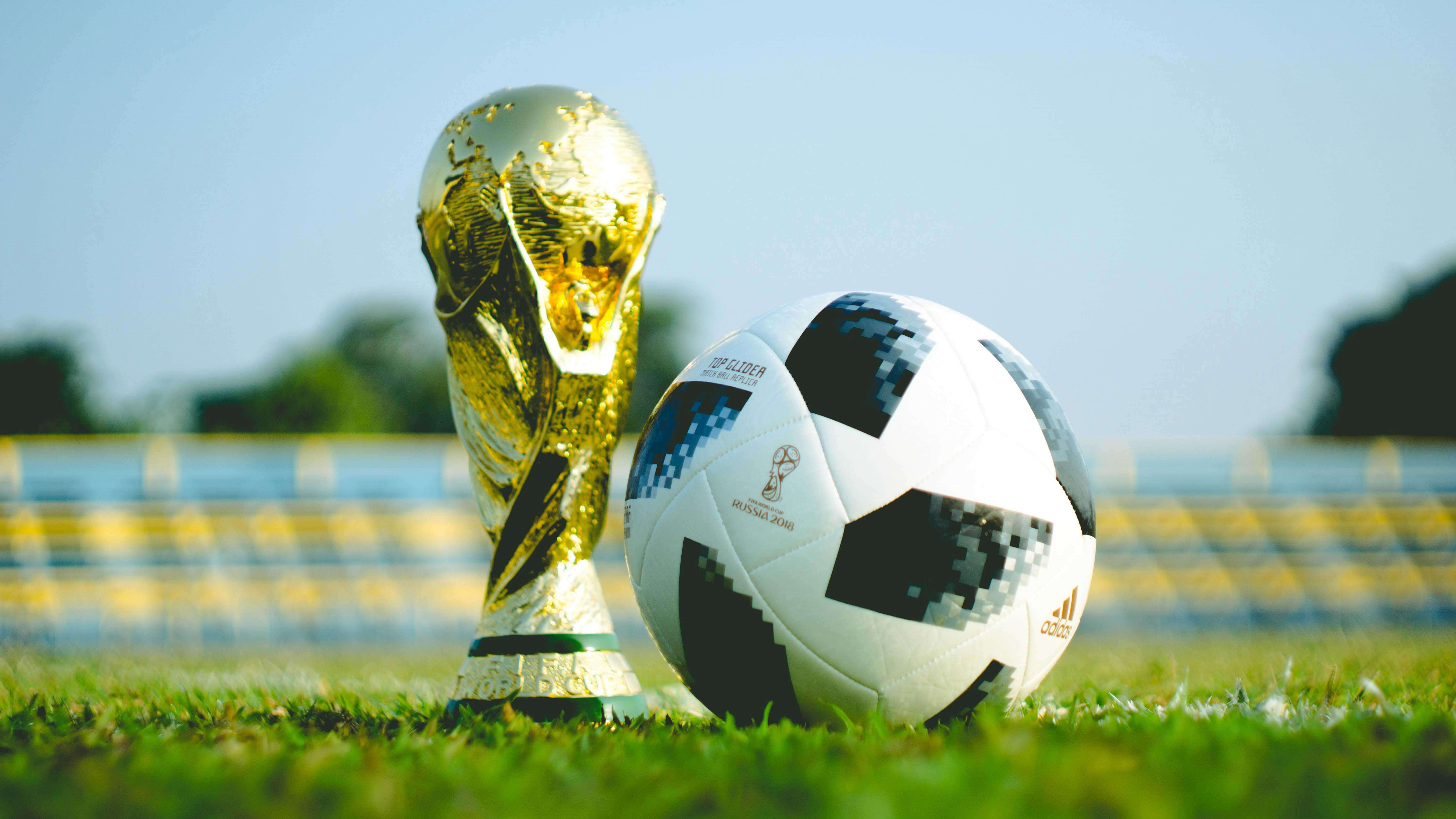 Gramado do campo de futebol com a taã da Copa do Mundo no chão ao lado de uma bola de futebol