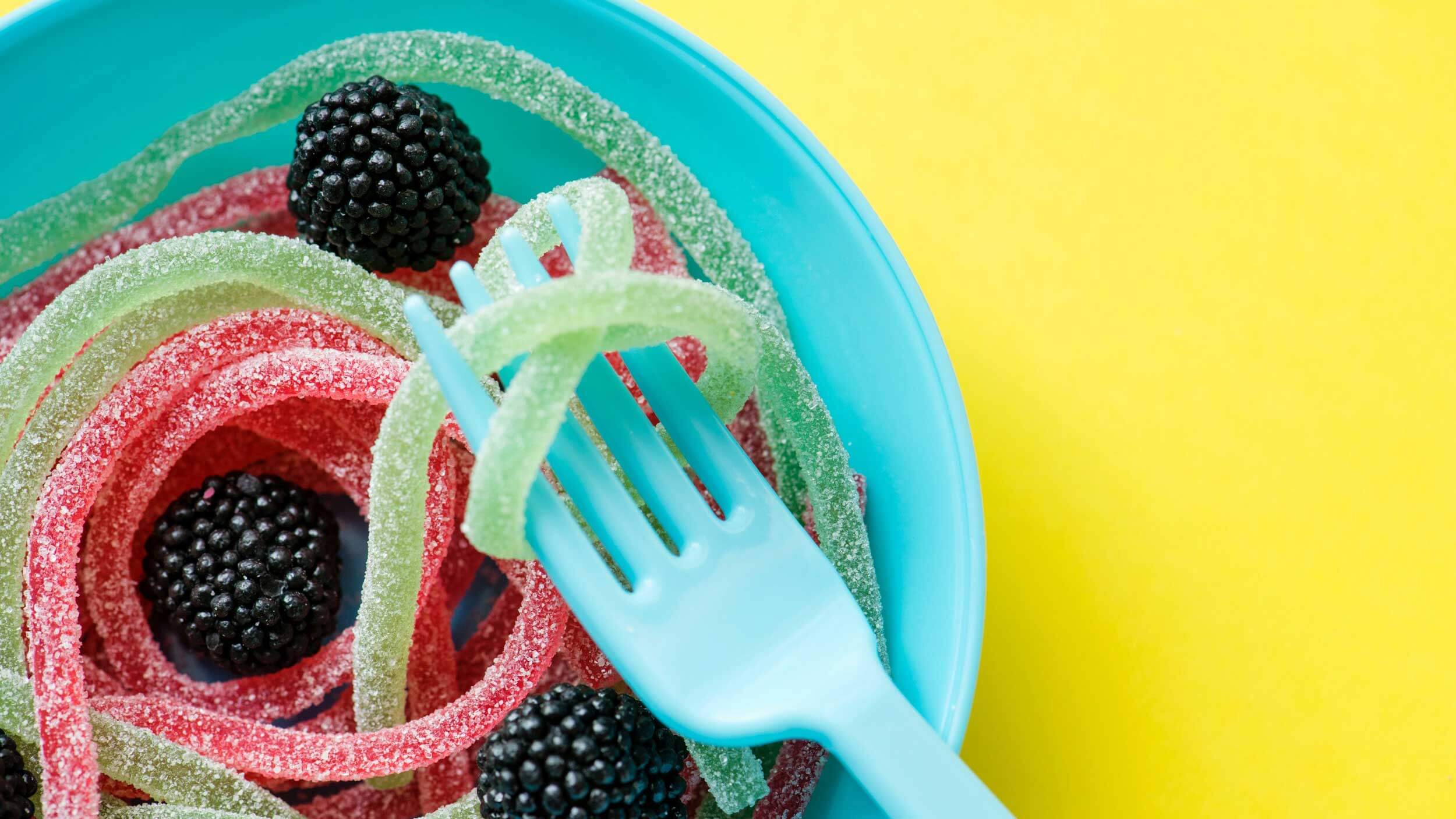 Prato azul com balas de gelatina em formato de amora e minhocas com um garfo levantando uma como se fosse um macarrão