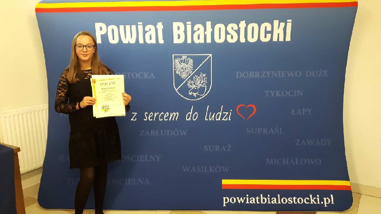Monika z dyplomem na te niebieskiego plakatu Powiatu Białostockiego z herbem i hasłem z sercem do ludzi