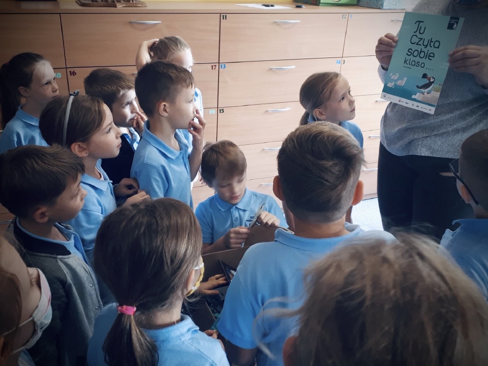 grupka dzieci, któym wychowawczyni pokazuje plakat programu Tu czyta sobie pierwsza klasa