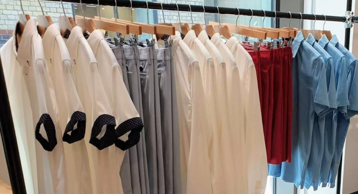 white and navy golf shirts, grey dress pants, white long sleeve dress shirt, red dress shorts and light blue polos hanging on a clothes rack in store.