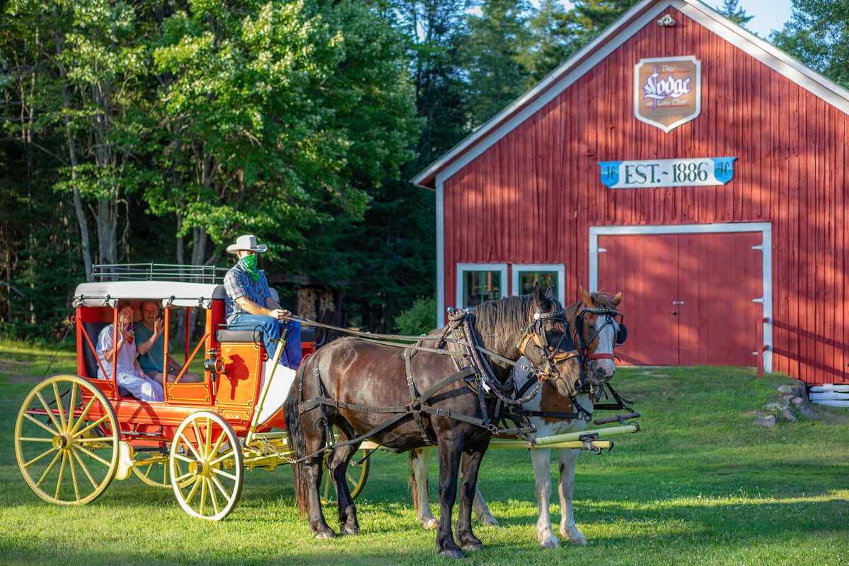 Guests sit in an 1850s Henderson Stagecoach pulled by draft horses in front of the original 1886 Stagecoach Barn at the Lake Clear Lodge & Retreat.