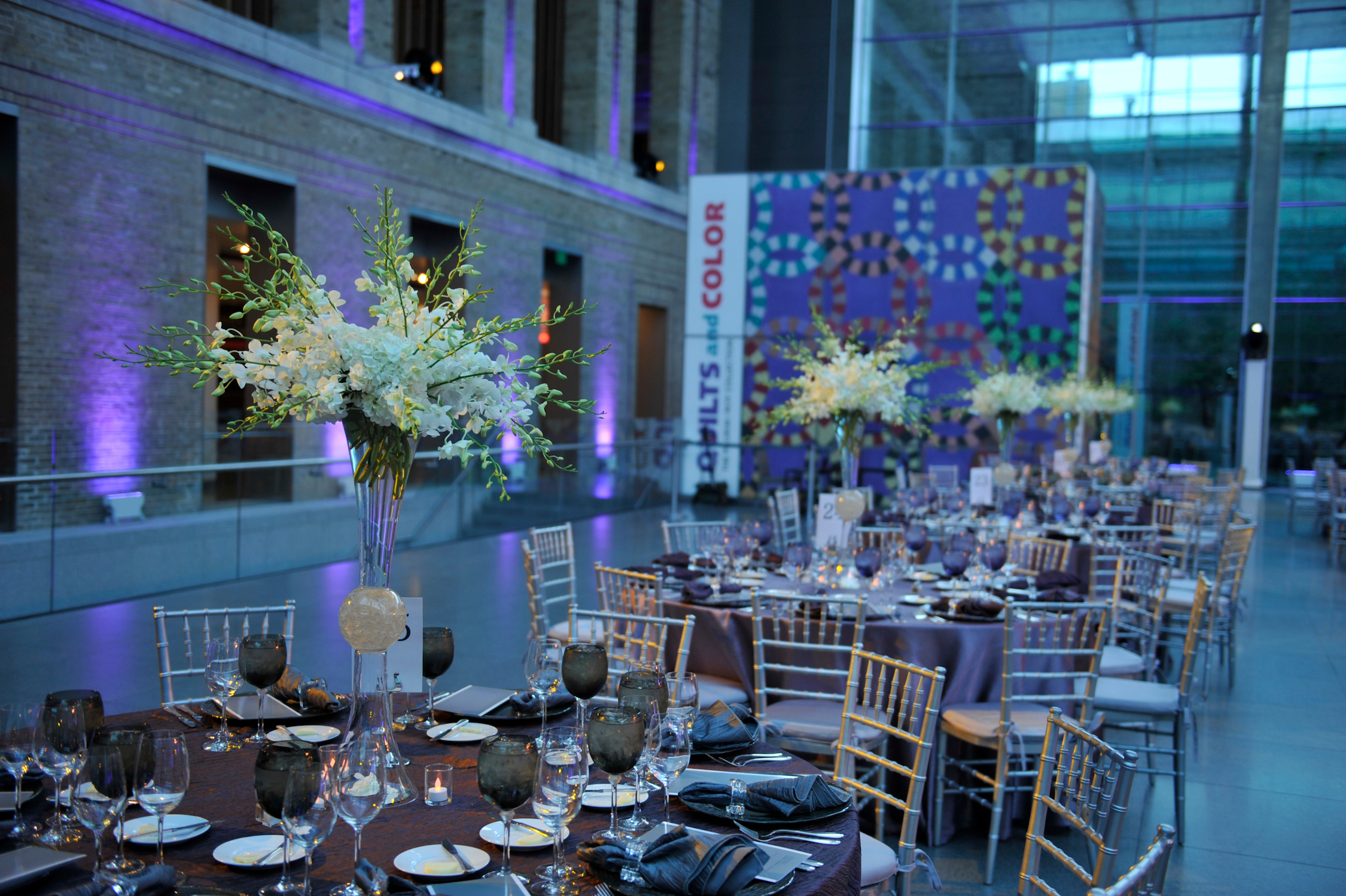 White floral centerpieces on purple tables with silver chairs in a vaulted, glass and stone room.