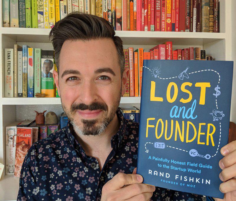 This is a picture of a man holding a book. He is the author of this book. This is a photo of Rand Fishkin, former CEO of Moz, a SaaS SEO product.