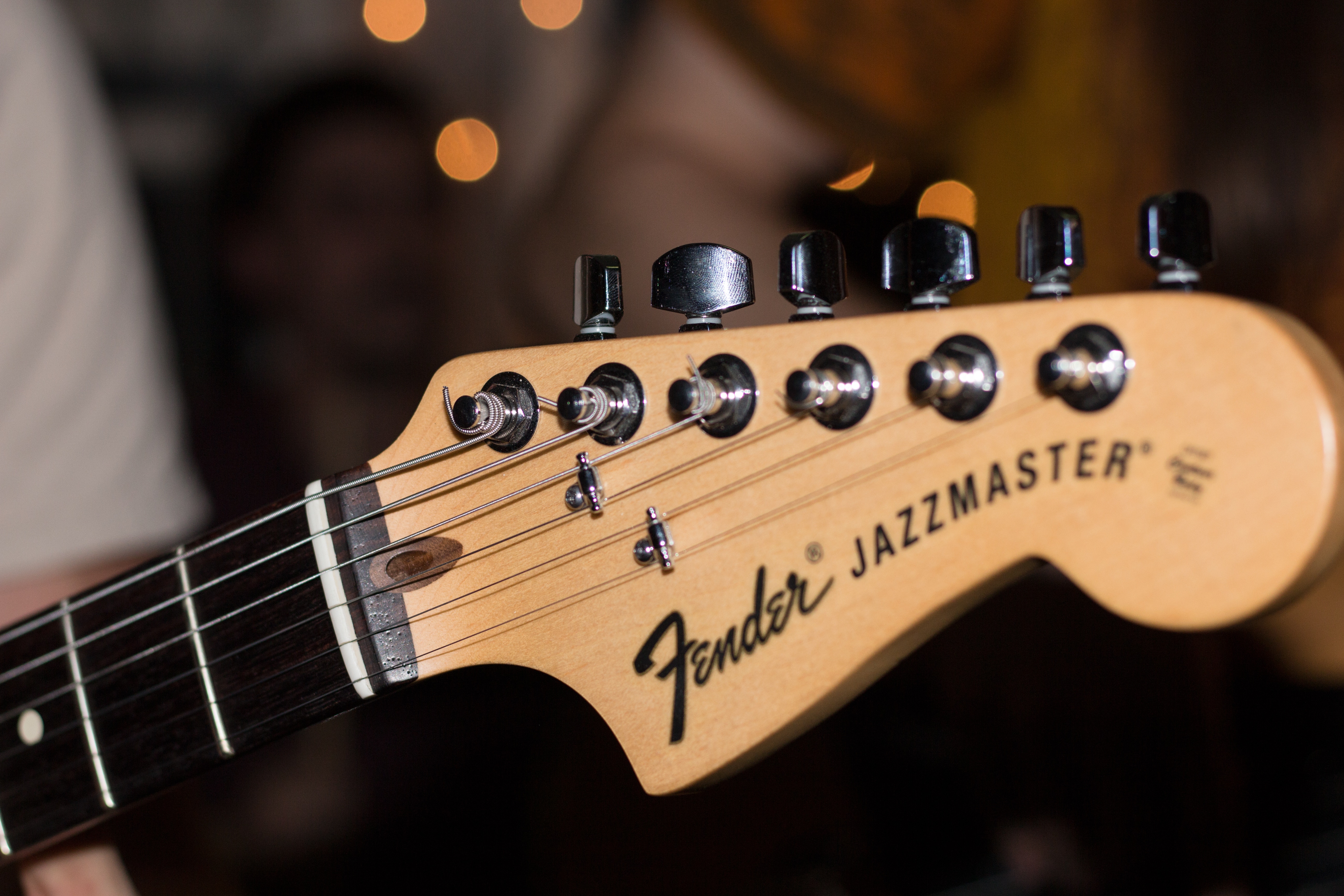 This picture is of a fender jazzmaster guitar handle with strings. It shows a person playing the guitar up close with a bokeh effect in the background.