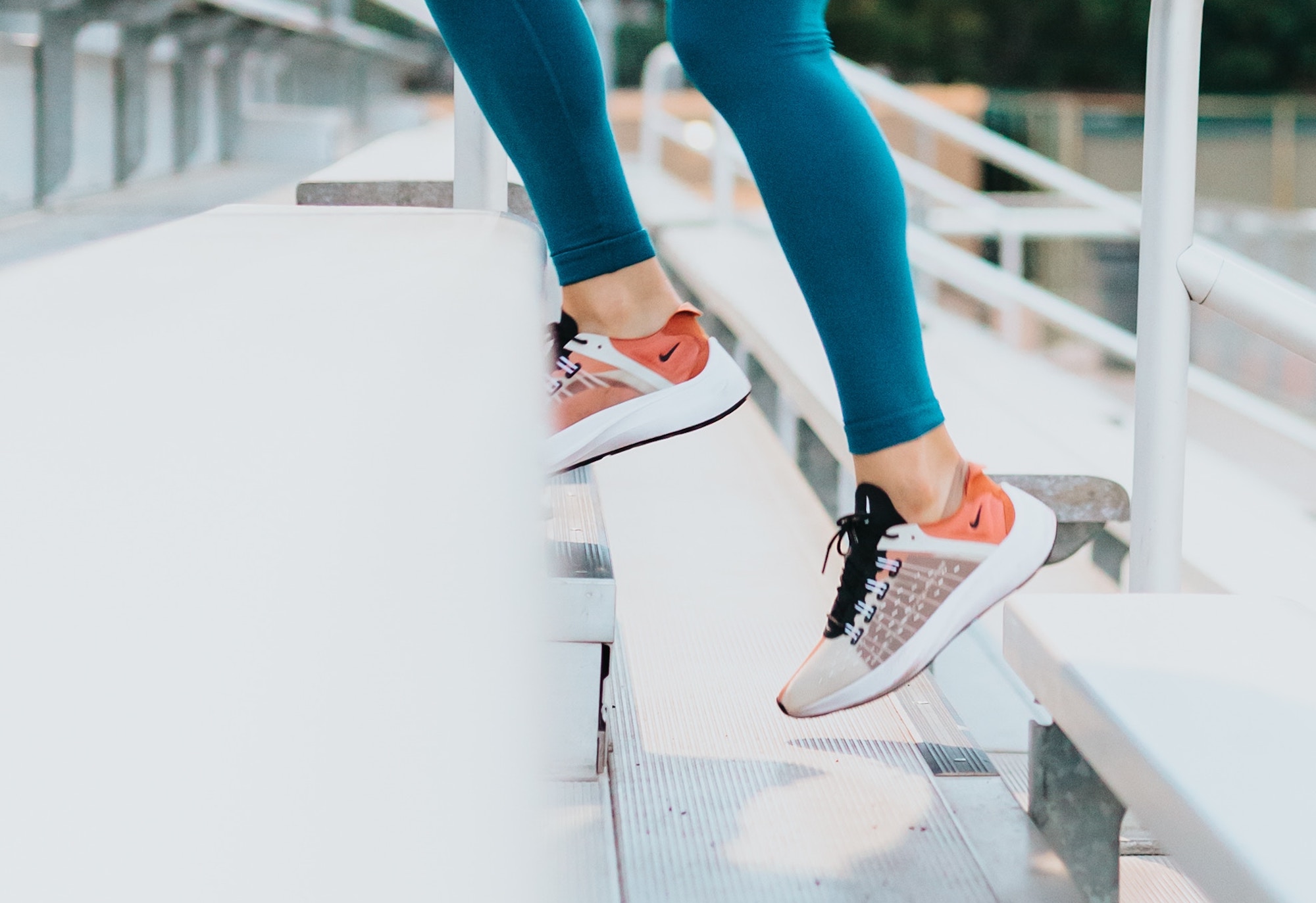 This picture shows a woman running up bleacher stairs wearing nike sneakers and teal leggings. This is a photo abaout fitness and performance footwear