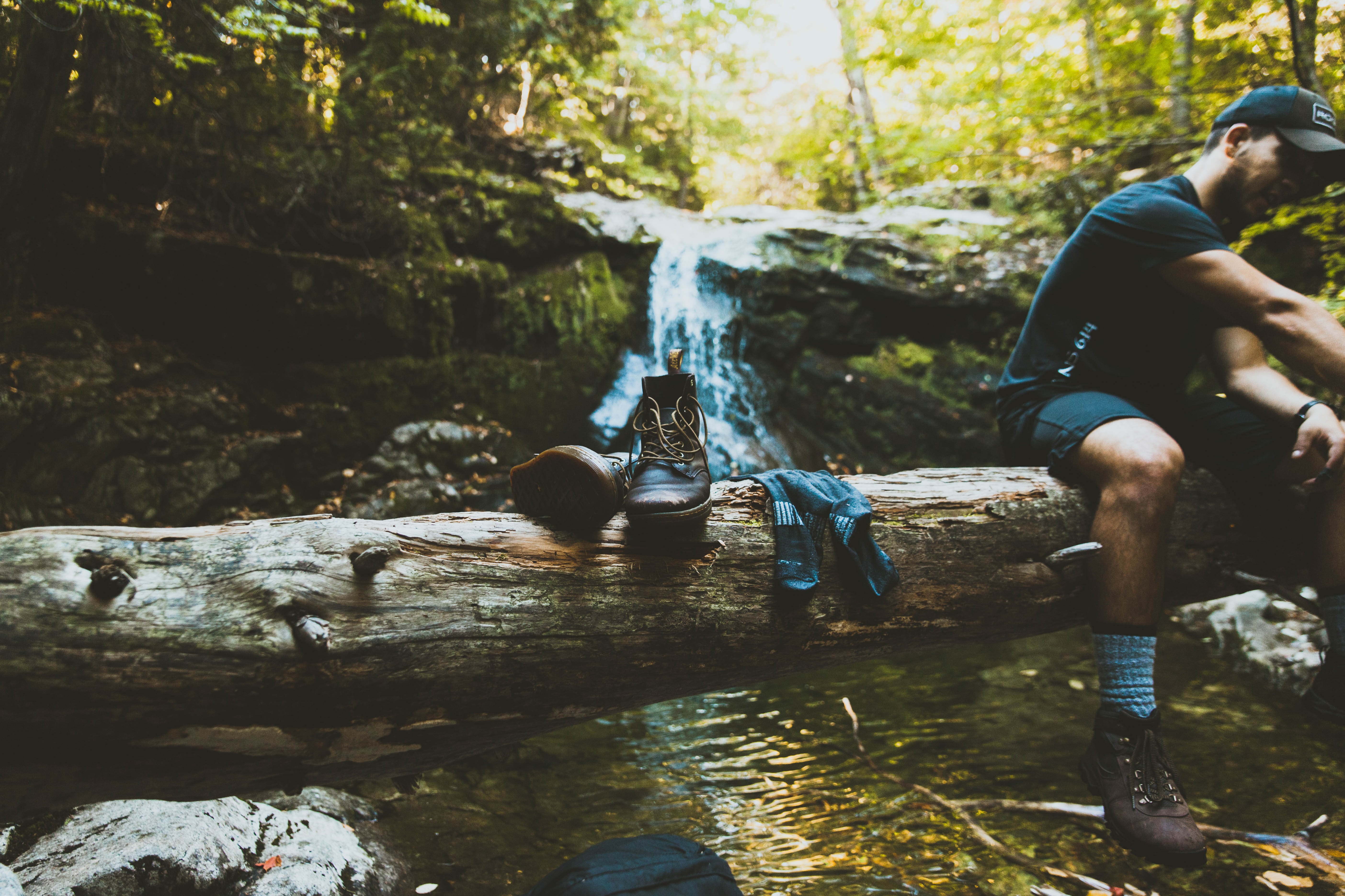 This is a picture of a man on hike, resting by a small waterfall. He is wearing boots with socks and sitting on a log.