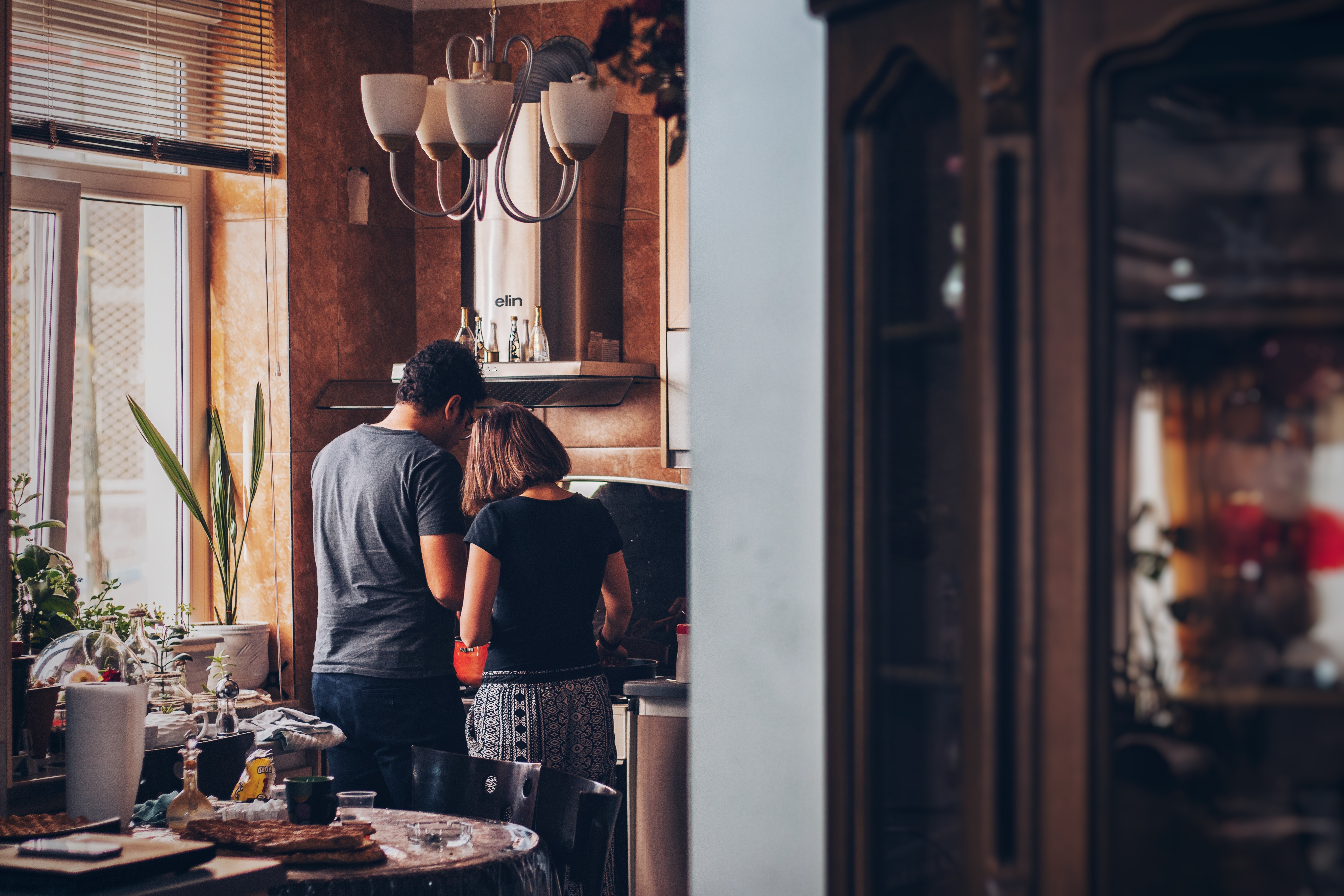 This is a picture of two people cooking in their kitchen. This is a photo of a couple cooking dinner by a window with stainless steel appliances.