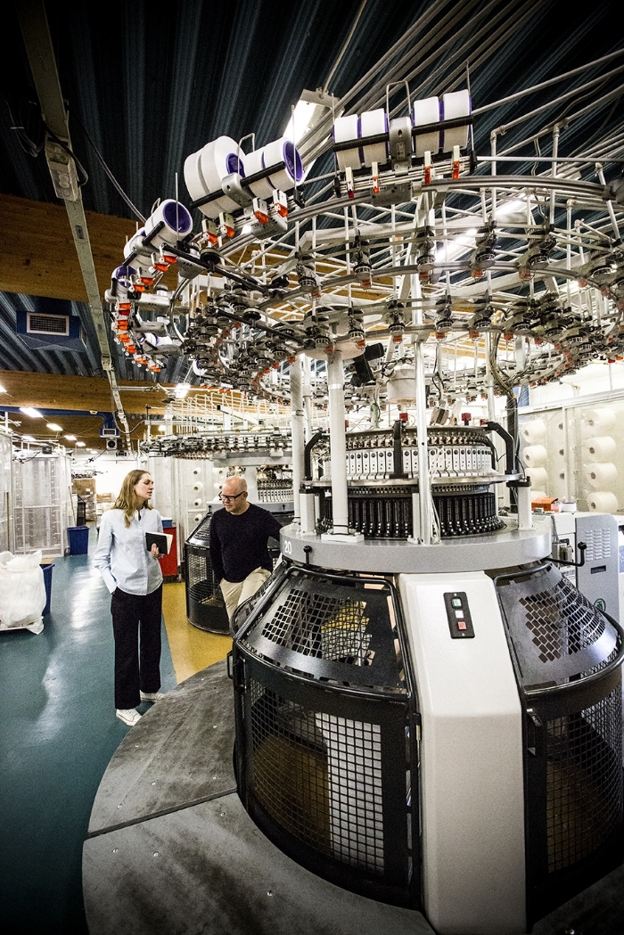 A woman, Lisa, and a man, Jonas Lorentzon, are standing by a huge, circular machine that towers over them. The machine has spools of thread connected to it, and is preparing to knit material.