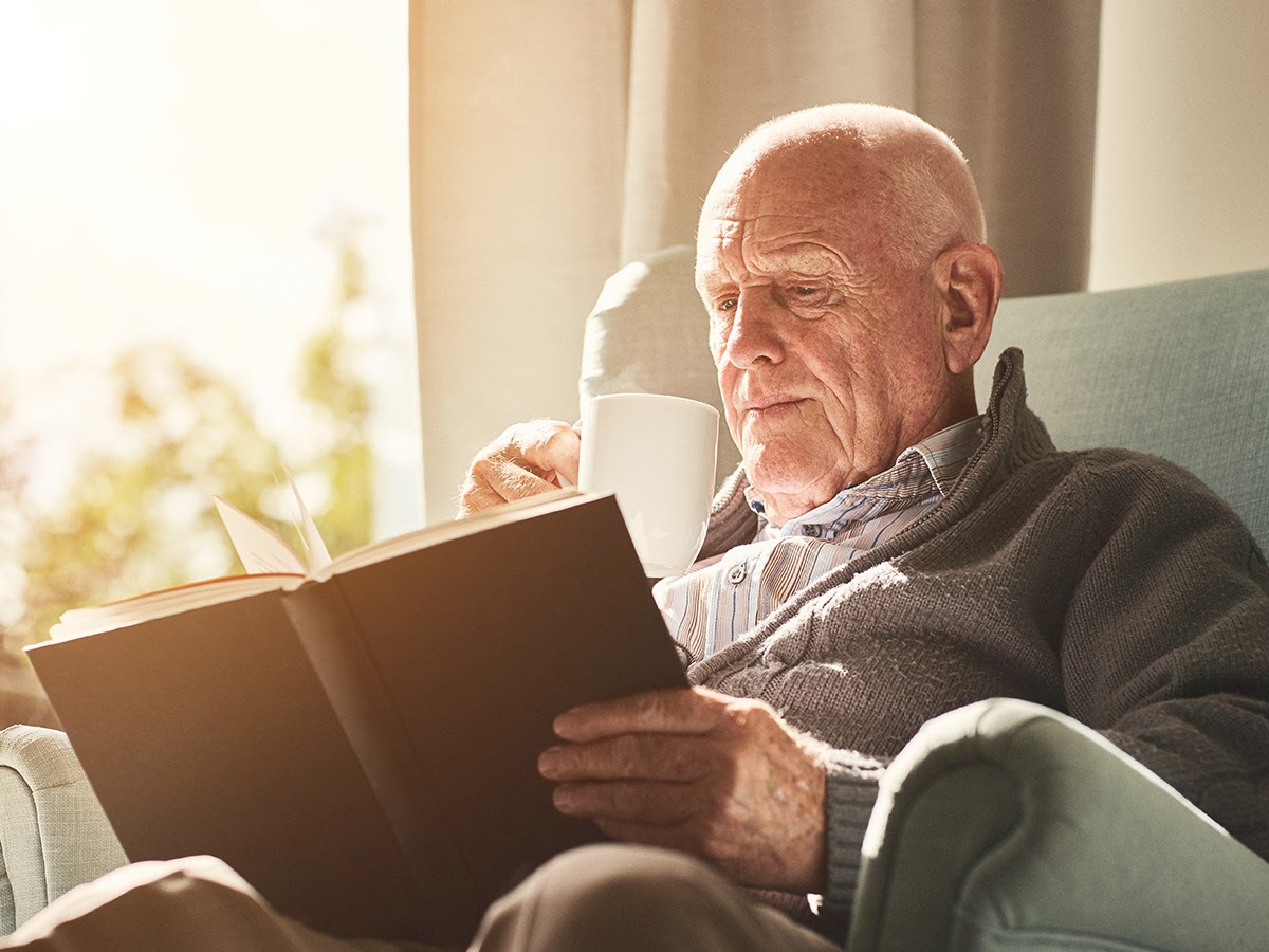 An elderly man sits in an armchair reading a book