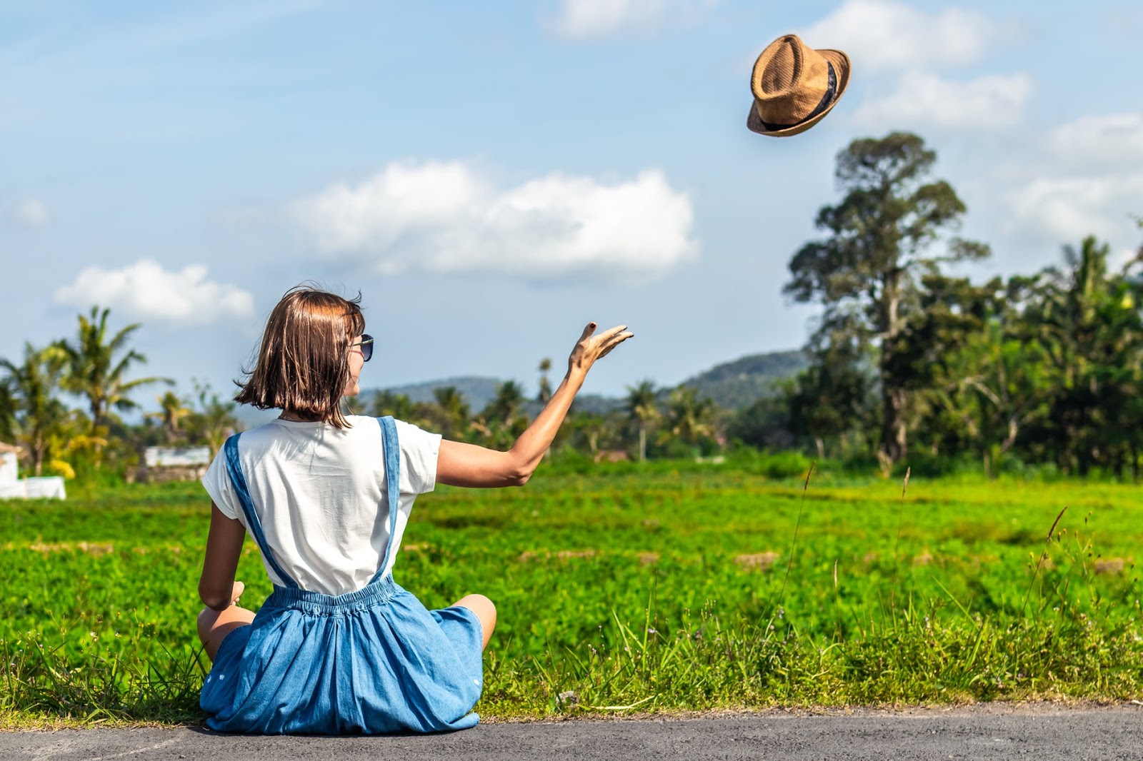 A girl in a blue skirt sits cross legged in a field and throws her hat into the air with a sense of freedom