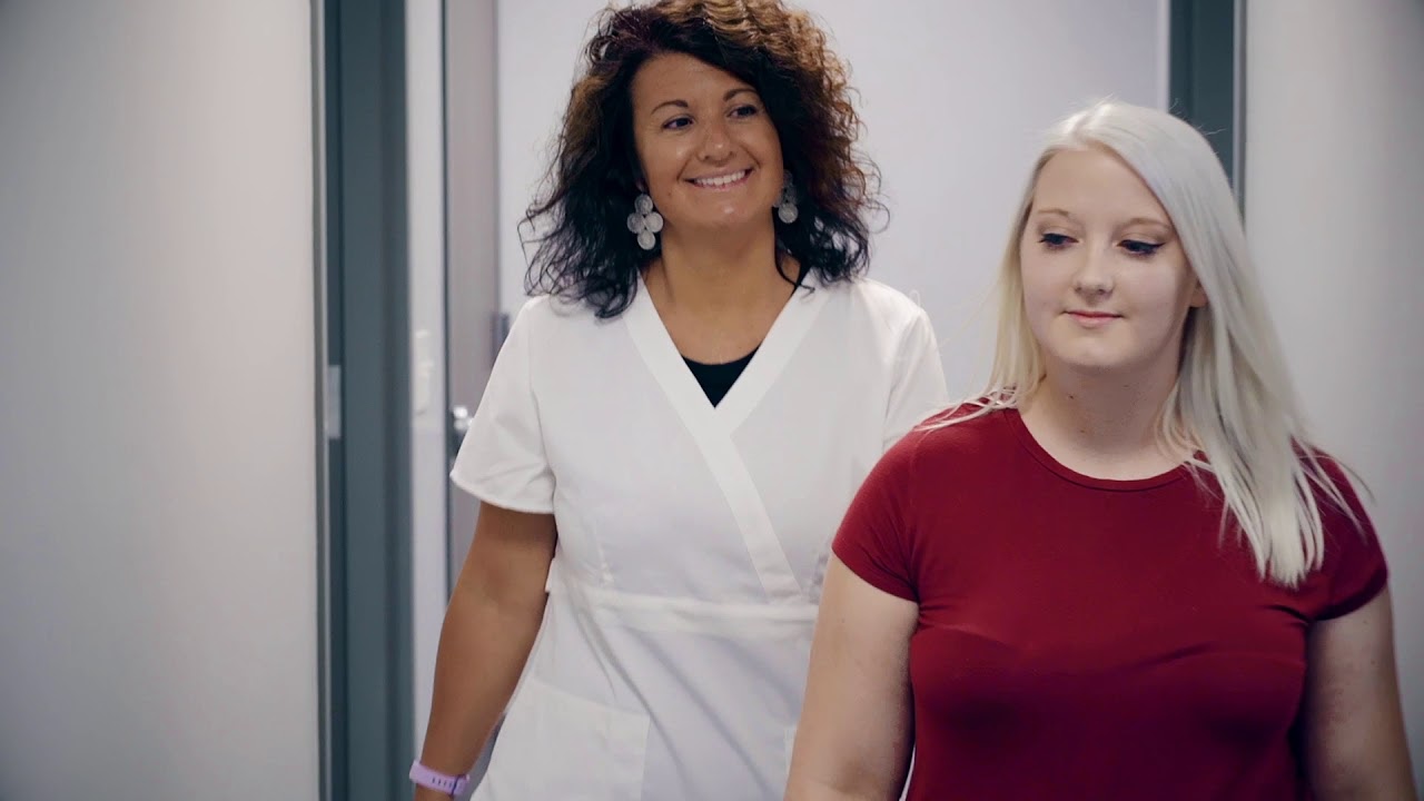 A health care professional leading a blond haired woman down the hall of a doctor's office.